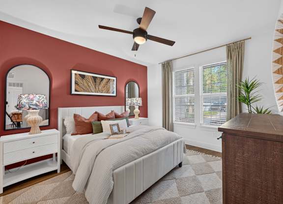 A bedroom with a large bed, a dresser, and a ceiling fan, the wall behind the bed is painted a soft red with a mirror above the bedside table