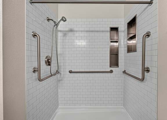 A white tiled shower with a glass door and a metal grab bar. Steel fixtures updated shower with  storage.