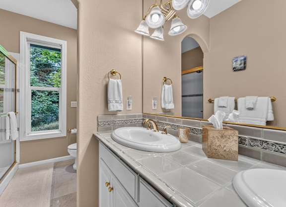A bathroom with a sink, mirror, and a window. White towels, light fixtures around the mirror, double sink vanity. Glass shower doors fora standing shower.