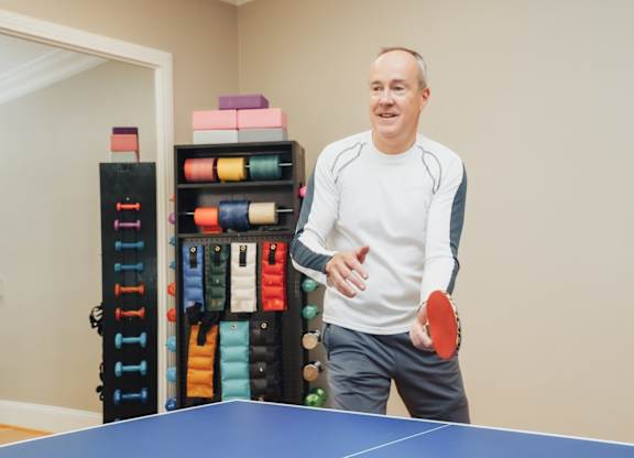A man is playing ping pong in a room with a shelf of sports balls in the background.