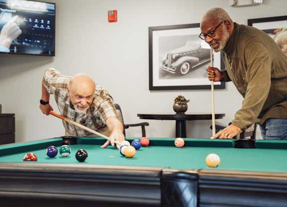 Two men playing pool in a room with a TV and a vintage car poster on the wall.