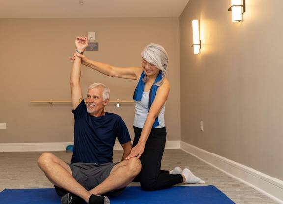A woman is stretching the arm of a man who is sitting on a mat.