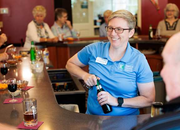 A woman in a blue shirt is pouring a drink for a man at a bar.