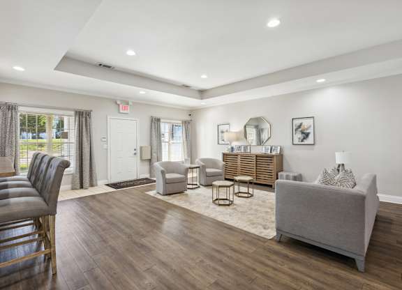 A well-lit, spacious living room with a grey sofa, wooden chairs, and a rug.