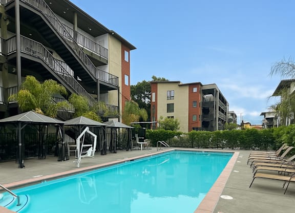 A swimming pool surrounded by lounge chairs and umbrellas in front of apartment buildings.