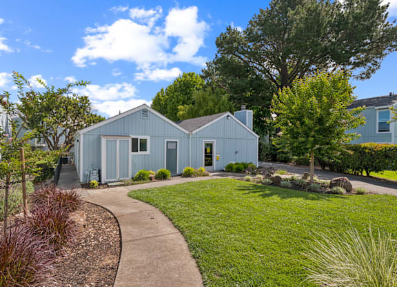 A blue house with a white door and windows is surrounded by greenery and a clear blue sky.