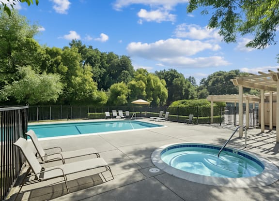 A pool with a hot tub and chairs in the foreground with trees in the background.