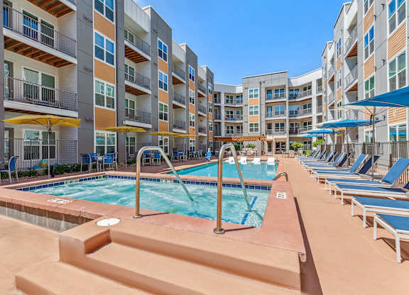 A large outdoor pool surrounded by lounge chairs and umbrellas in front of apartment buildings.
