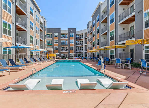 A large swimming pool surrounded by lounge chairs and umbrellas in front of apartment buildings.