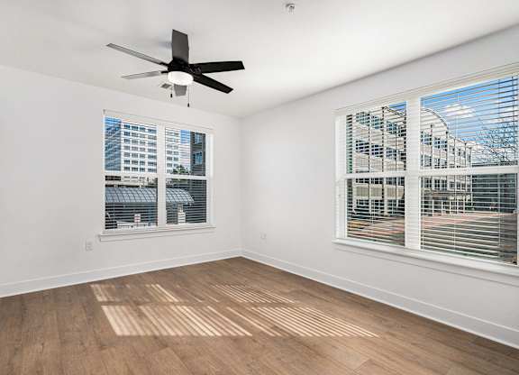 A room with a ceiling fan and two windows overlooking a cityscape.
