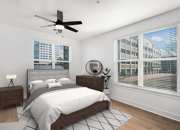 A bedroom with a large bed, a ceiling fan, and a view of a cityscape through the window.