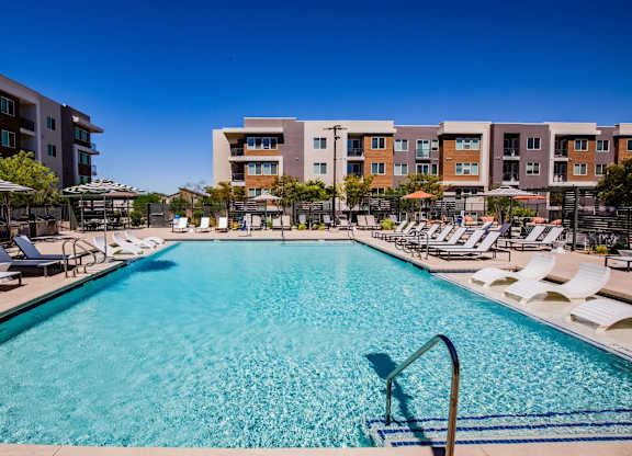 A large swimming pool surrounded by sun loungers and umbrellas in front of apartment buildings.
