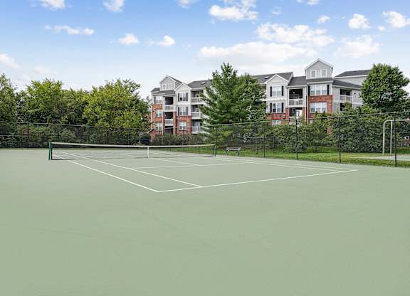 A tennis court with a green surface and white lines, surrounded by a fence and with apartment buildings in the background.