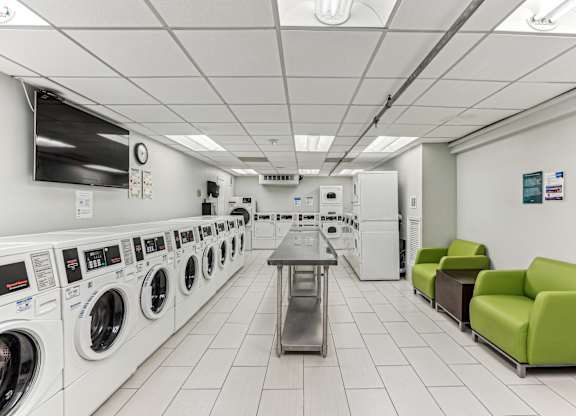 A laundromat with a row of washers and dryers on the left and a table and two chairs on the right.