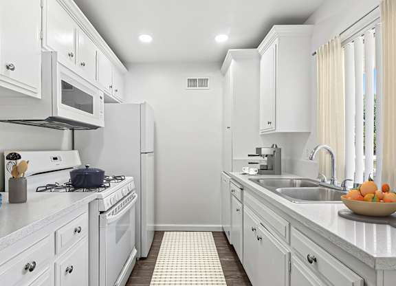 Kitchen in 955 Via Zapata apartment home with wood-style plank flooring, white cabinetry, white appliances, and a view of the stunning trees throughout the community.