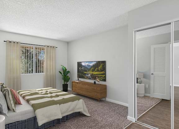 Oversized bedroom in 955 Via Zapata apartment home. Entryway inside the bedroom has wood-style plank flooring, a large closet with mirrors and a hallway on the right leading to a private vanity area and private bathroom.  Left side of room has plush carpeted flooring and large windows allowing for gorgeous natural lighting.