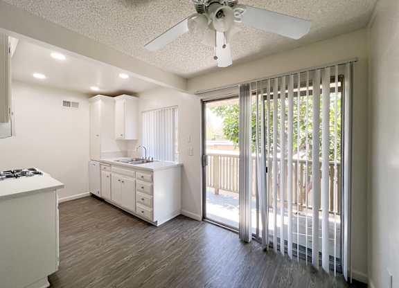 Dining nook & kitchen in 955 Via Zapata apartment home with wood-style plank flooring, white cabinetry, overhead ceiling fan and sliding doors leading to private balcony shaded by gorgeous trees.