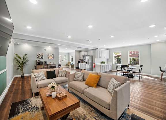 Spacious community clubhouse with lounge area including two sofas, an area rug, coffee table and tv. Open concept layout leads to two dining areas and a kitchenette.