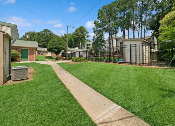 A grassy area with a concrete walkway in the foreground and a fence.