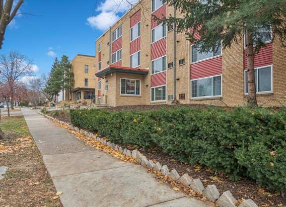 A sidewalk runs alongside a row of bushes and trees in front of a red brick apartment building.