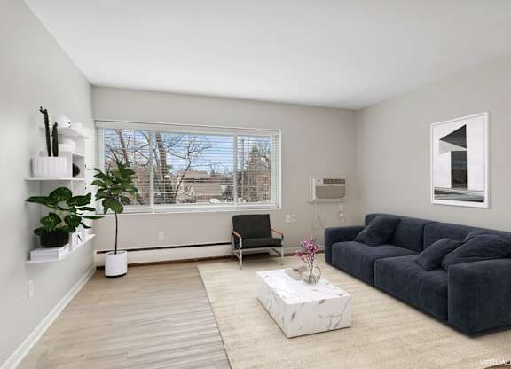 A living room with a grey sofa, a black chair, a white coffee table, and a window with a view of trees and a building.