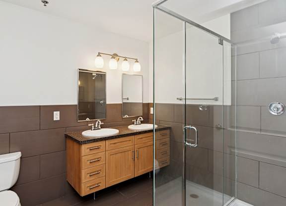 A bathroom with a white toilet, a wooden vanity with two sinks, and a glass shower stall.