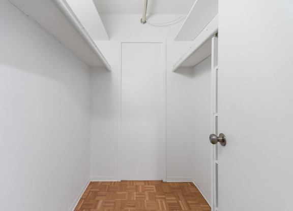 A walk-in closet with white door with a silver handle is in a white room with a wooden floor.