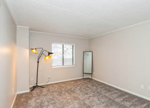 A bedroom with a carpeted floor, a window with blinds, a lamp with a yellow shade, and a mirror.