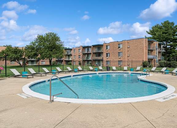 A swimming pool surrounded by lounge chairs and trees in front of a brick apartment building.