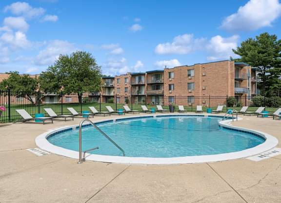 A swimming pool surrounded by lounge chairs and trees in front of a brick apartment building.
