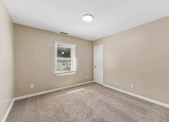 A room with a carpeted floor, a window with blinds, and a white door.