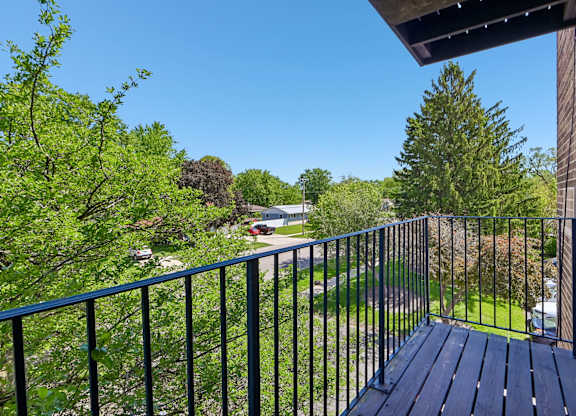 A balcony with a black railing and a wooden floor overlooks a green lawn and trees.