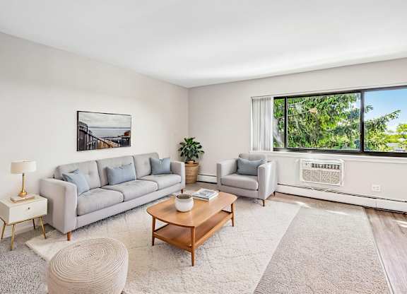 A living room with a grey couch, a wooden coffee table, a white rug, a white wall, a window with a view of trees, and a painting hanging on the wall.