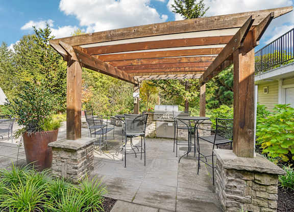 A wooden pergola over a patio with chairs and tables with a bbq.