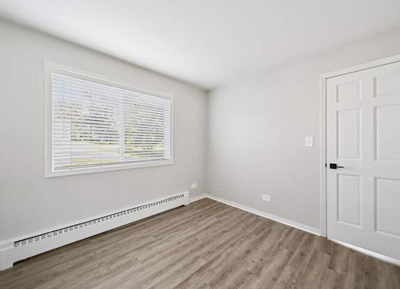 A room with a white door, window with blinds, and a radiator on the wall.
