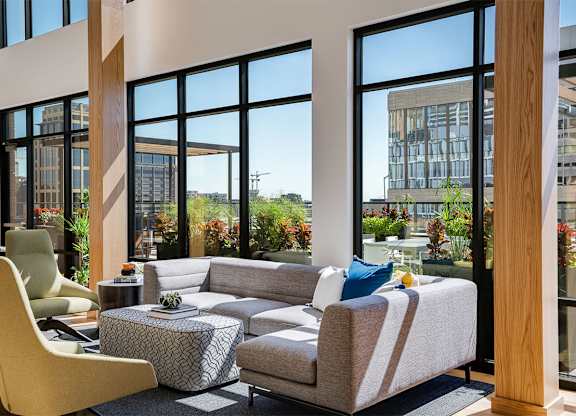 Front lounge with a yellow chair and a grey couch and large windows in the background at Sora Apartments, Minneapolis, MN