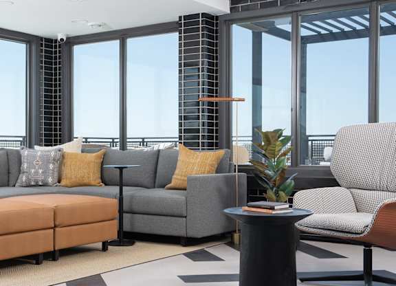 A living room with a grey couch, a brown ottoman, and a black table with a plant on it.