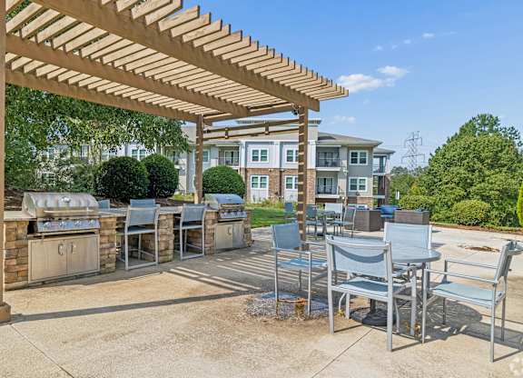 A wooden pergola with metal chairs is in front of a building.