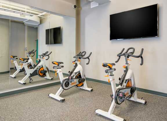 Three stationary bikes are lined up in a row in a room with a wall-mounted TV.