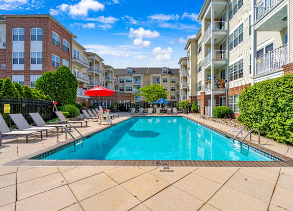 A pool surrounded by chairs and umbrellas with apartment buildings in the background.