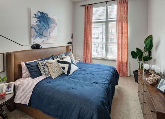 A bedroom with a blue bedspread and pillows, a wooden headboard, and a window with orange curtains at Regatta Sloans Lake Apartments, Denver, 80204