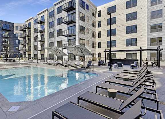 A large pool surrounded by sun loungers and umbrellas in front of apartment buildings.