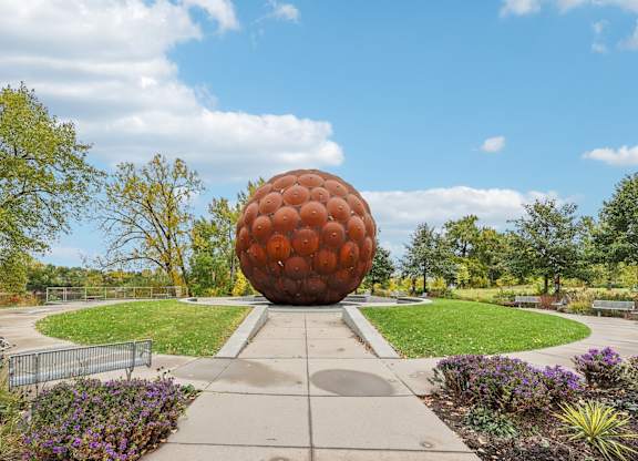A large orange sphere sculpture is the centerpiece of a park with a walkway leading to it.