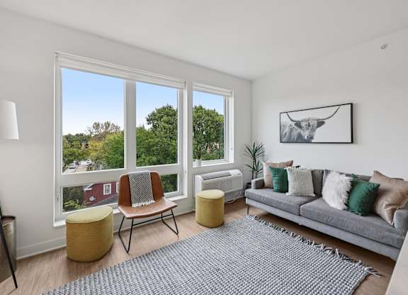 A living room with a grey couch, a yellow ottoman, a grey rug, a chair, a floor lamp, and a picture on the wall.