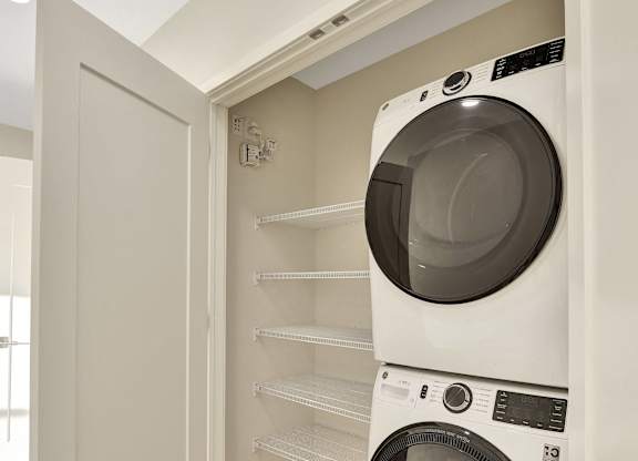 A white washing machine is in a small space between a wall and a shelf.