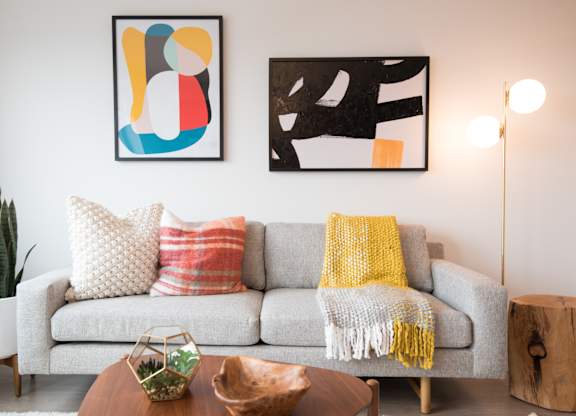 A living room with a grey couch, a yellow throw, and two pieces of abstract art.