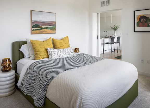 A bedroom with a bed, a painting on the wall, and a dining area in the background.