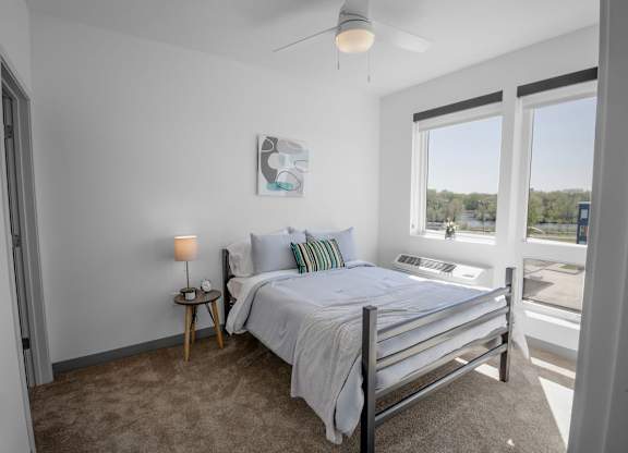 A bedroom with a bed, a nightstand with a lamp, and a window overlooking a parking lot.