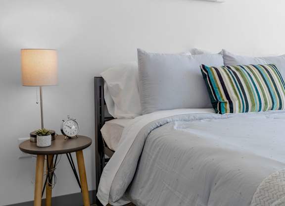 A bed with a grey comforter and striped pillow beside a table with a lamp and a small plant.