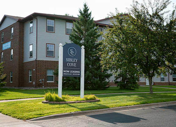 A sign for Sibley Cove, a now leasing apartment complex, stands in front of a brick building.